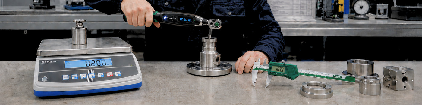 Técnico usando llave de torque digital junto a balanza de precisión, calibre vernier y piezas metálicas mecanizadas sobre banco de trabajo